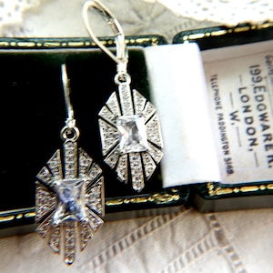 May include: Silver Art Deco earrings featuring a central rectangular stone and surrounding smaller stones. The earrings are presented in a dark green jewellery box with gold trim. The box has text that reads "199, EDGWARE R., LONDON, W."