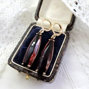 Puede incluir: Un par de pendientes de oro con gotas de piedras preciosas rojas y negras en forma de lágrima. Los pendientes se muestran en una caja de joyería forrada de terciopelo.