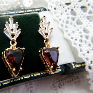 May include: A pair of gold-toned earrings with a heart-shaped, dark red gemstone pendant. The earrings have a gold frame with small, round accents and a cluster of clear, rectangular crystals at the top. The earrings are displayed on a dark green surface.