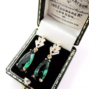 May include: Gold-plated drop earrings featuring emerald green pear-shaped gems and clear baguette-cut details. The earrings are presented in a black and white jewellery box with gold edging.