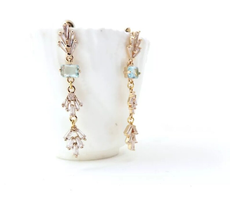 May include: Gold dangle earrings with a light blue gemstone and clear crystal accents.
