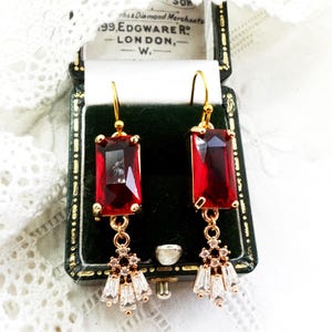 May include: Gold-coloured drop earrings with large, rectangular, red gemstones. Each earring has a cluster of smaller, clear, and gold-coloured crystals below the red stone. The earrings are displayed in a vintage jewellery box.