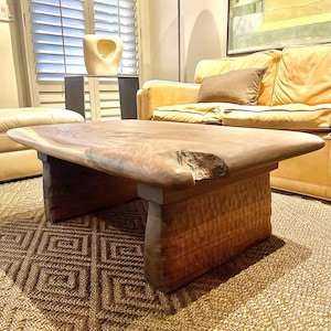 May include: A handcrafted wooden coffee table with a natural, organic shape. The table features a light brown, live-edge top and textured, carved legs. The table is set on a woven rug in a living room setting.