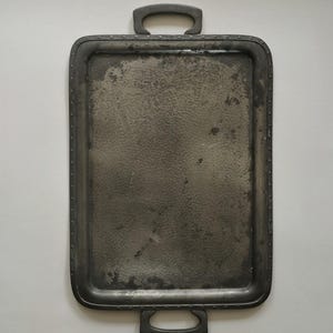 May include: A rectangular metal serving tray with a dark, weathered finish and a hammered texture on the surface. The tray features two sturdy handles on opposite ends and a decorative border with small, evenly spaced indentations along the rim.