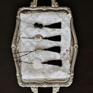 May include: A silver serving tray holds four necklaces with black tassels. The necklaces have different pendants: black beads, an oval silver pendant, a silver cylinder, and a white circular pendant. The necklaces rest on a white lace doily.