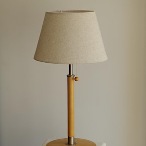 May include: A wooden table lamp with a beige fabric shade. The lamp has a light wood base and a cylindrical wooden stem with a metal accent. The shade is conical and the lamp is on a white surface.