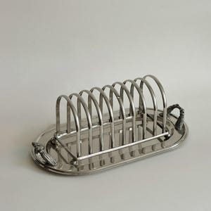 May include: A silver-toned metal toast rack featuring an oval base tray with a polished finish. The rack consists of seven vertical arched dividers for holding slices of bread, and the tray includes two decorative textured handles on each end.