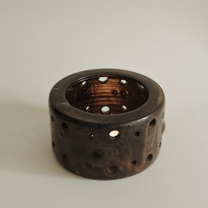 May include: A cylindrical ceramic candle holder with a dark, mottled brown glaze. The exterior and interior walls feature numerous circular cutouts of varying sizes, allowing light to pass through the thick, textured clay structure.