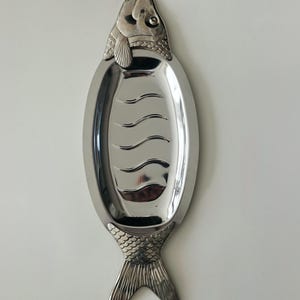 Könnte beinhalten: Eine silberfarbene Servierplatte aus Metall in Fischform. Die Platte verfügt über eine polierte, glatte ovale Mitte mit geprägten Wellenlinien, während Kopf- und Schwanzbereich mit detaillierten Schuppenmustern und ausgeprägten Flossen texturiert sind.