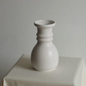 Op de afbeelding: Een matwitte keramische vaas met een ronde voet, een smalle hals en een uitlopende rand. De vaas heeft een glad, effen oppervlak met een opvallend horizontaal ribbeldetail rond de hals, wat zorgt voor een gebalanceerd en symmetrisch silhouet.