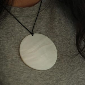 Könnte beinhalten: Ein großer, kreisförmiger, cremeweißer Anhänger mit einer glatten, perlmuttartigen Oberflächenstruktur. Der Anhänger hängt an einer dünnen, schwarzen Kordel, die durch ein kleines Loch in der oberen Mitte der Scheibe verläuft und eine schlichte Kette bildet.