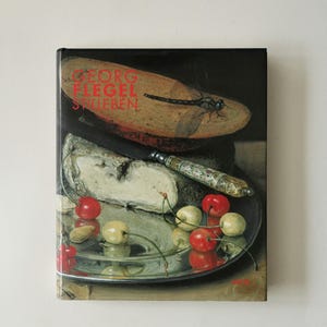 May include: A hardback book titled Georg Flegel Stilleben featuring a still life painting on the cover. The artwork depicts a silver platter with cherries, a wedge of cheese, a knife with an ornate handle, and a dragonfly resting on bread.