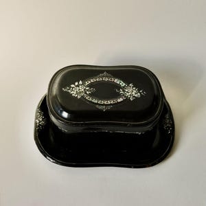 May include: A black, oval-shaped trinket box with a matching tray. The box lid features a decorative oval design with floral and pearl accents. The tray has a scalloped edge. The box and tray are made of a glossy material.