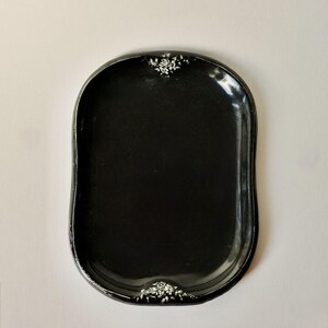 May include: A black, oval-shaped tray with a glossy finish. The tray has a slightly raised edge and is decorated with white floral accents at the top and bottom. The tray is empty and sits on a white background.