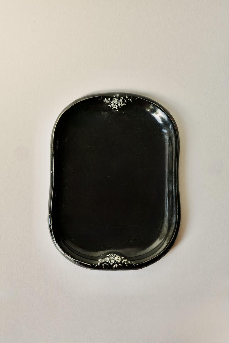 May include: A black, oval-shaped tray with a glossy finish. The tray has a slightly raised edge and is decorated with white floral accents at the top and bottom. The tray is empty and sits on a white background.