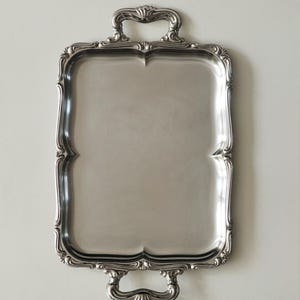 May include: A rectangular silver-toned serving tray with a smooth, reflective surface and a raised, ornate border. The tray features two integrated handles at the top and bottom, each decorated with detailed scrollwork and shell-like motifs.