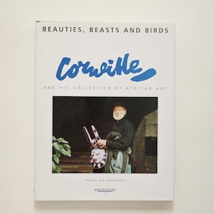 以下が含まれることがあります： Beauties, Beasts and Birds, Cornille and his collection of African Art（Ronald A.R. Kerkhoven著）というタイトルの白いソフトカバーの本。表紙には、白いひげを生やした人物がマスクを持っている写真と、Jaski Art Galleryというテキストが印刷されています。