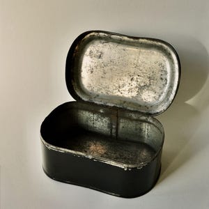 May include: An open, rectangular black tin with a hinged lid. The interior of the tin and lid are silver with signs of wear. The tin is set against a white background.