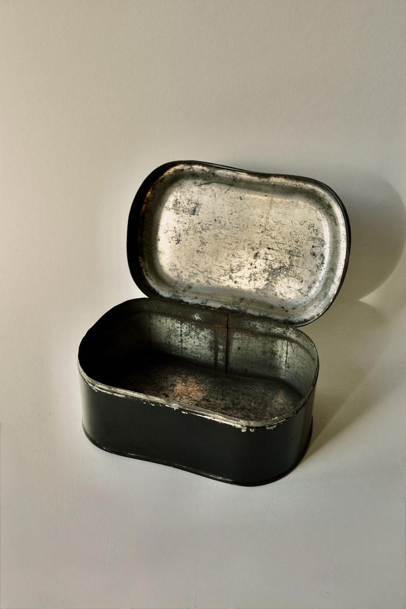 May include: An open, rectangular black tin with a hinged lid. The interior of the tin and lid are silver with signs of wear. The tin is set against a white background.