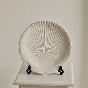 Könnte beinhalten: Ein weißer, muschelförmiger Dekoteller mit geripptem Design. Der Teller wird auf einem weißen Sockel mit schwarzen Stützen präsentiert, wodurch ein Küsten- oder Maritim-Thema entsteht. Das Design des Tellers ähnelt einem Fächer oder einer Muschel.