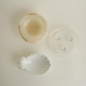 Op de afbeelding: Een set van drie kleine schaaltjes, bestaande uit een ronde beige stenen kom met natuurlijke aders, een doorzichtig glazen bordje met drie kleine ronde voetjes aan de onderkant en een wit keramieken schaaltje in de vorm van een schelp.