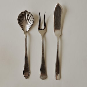 Pode incluir: Três talheres de servir prateados dispostos sobre uma superfície branca. O conjunto inclui uma colher com uma tigela em forma de concha, um garfo de dois dentes e uma faca de manteiga. Os utensílios têm cabos decorativos.