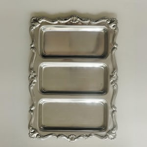 Könnte beinhalten: Ein rechteckiges Serviertablett aus silberfarbenem Metall mit drei separaten, vertieften Fächern. Das Tablett verfügt über einen verzierten, dekorativen Rand mit erhabenen Schnörkeln und muschelartigen Motiven an den Kanten.