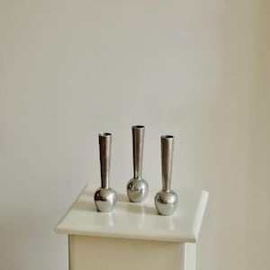 Puede incluir: Tres pequeños jarrones plateados con base redondeada y cuello largo y delgado. Los jarrones se exhiben sobre una superficie blanca sobre un fondo neutro. Los jarrones son de metal y tienen un acabado brillante.