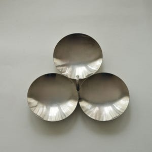 May include: A silver-toned metal serving dish featuring three circular, bowl-shaped compartments arranged in a clover-like formation. The bowls are connected at the center by a small, textured metal ring, creating a unified, symmetrical piece.