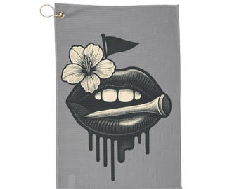 May include: Gray golf towel featuring a black and white illustration of lips with a golf tee and a flower. The lips have a dripping effect. A small flag is above the lips. The towel has a gold-colored hook.