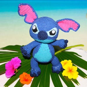 Peut inclure: Poupée Stitch bleue au crochet, avec des détails roses sur les oreilles, posée sur une feuille verte. La scène comprend des fleurs colorées et un fond de plage avec des palmiers et un ciel bleu.