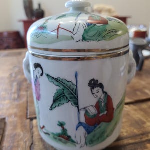 Può includere: Un vaso in porcellana bianca con un coperchio, decorato con scene dipinte a mano di persone in un paesaggio. Il vaso ha due manici sui lati e un bordo dorato intorno alla parte superiore.