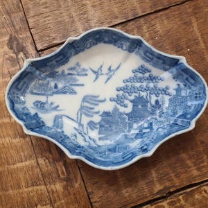 May include: A blue and white porcelain dish with a detailed landscape scene. The dish has a scalloped edge and features a traditional Asian design with trees, buildings, and birds. The color palette is primarily blue and white.