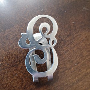 May include: A silver-colored money clip with an ornate, stylized letter "E" design. The clip features a curved, openwork pattern and a hinged base for securing bills. The clip is made of a shiny metal and rests on a brown surface.