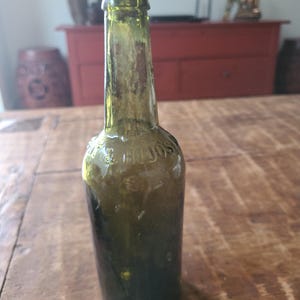 Könnte beinhalten: Eine dunkelgrüne Glasflasche mit abgerundetem Oberteil und leicht ausgestelltem Boden. Die Flasche ist alt und hat ein trübes Aussehen.