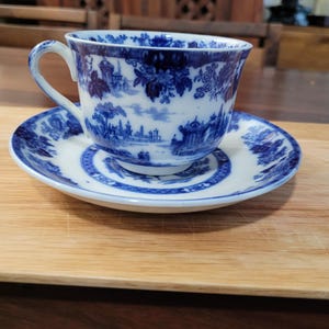 Puede incluir: Un juego de taza y platillo de porcelana azul y blanco con un diseño floral y de paisaje. La taza tiene un asa y el platillo tiene un borde festoneado.