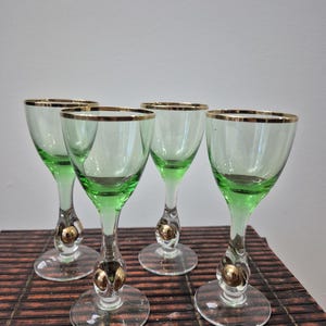May include: Four vintage green cordial glasses with gold rims and decorative gold accents on the stems. The glasses have a clear base and are arranged on a brown surface.