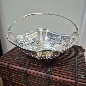 May include: A silver-colored metal basket with a polished finish and a high, arched handle. The bowl features an intricate, openwork filigree pattern along the sides, with a scalloped rim and a smooth, reflective interior base.