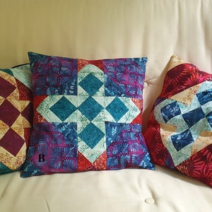 Könnte beinhalten: Drei dekorative Kissen mit einem geometrischen Muster in Rot-, Blau-, Grün- und Cremetönen. Die Kissen liegen auf einem hellbeigen Sofa.