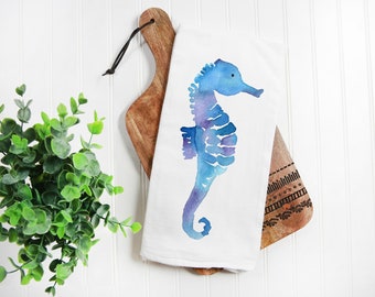 Puede incluir: Una toalla de cocina blanca con una ilustración de acuarela de un caballito de mar azul. La toalla está colgada en una tabla de cortar de madera con un patrón decorativo.