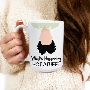 Op de afbeelding: Een witte keramische mok met een cartoonillustratie van een persoon ondersteboven met de tekst "What's Happening HOT STUFF?"