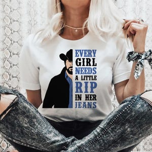 以下が含まれることがあります： カウボーイハットをかぶった男性のグラフィックと「Every girl needs a little rip in her jeans」というテキストがプリントされた白いTシャツ。