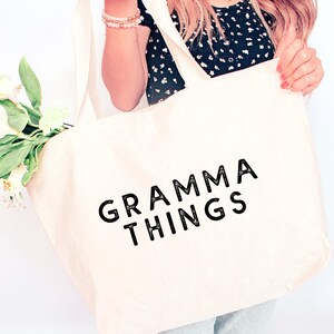 Könnte beinhalten: Eine cremefarbene Tragetasche mit dem schwarzen Aufdruck "GRAMMA THINGS". Die Tasche wird von einer Person gehalten, die ein schwarzes Oberteil mit Blumenmuster und hellblaue Jeans trägt. Die Tasche hat lange Träger.
