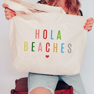 Op de afbeelding: Een witte canvas tote bag met de tekst "HOLA BEACHES" in regenboogkleuren. Een klein rood hartje bevindt zich onder de tekst.