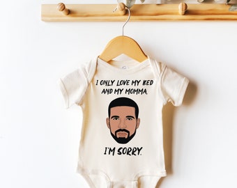 May include: A white baby onesie with a black illustration of Drake's face and the text "I only love my bed and my momma. I'm sorry."