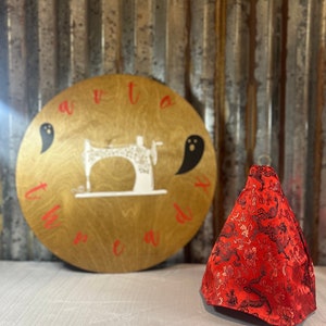 Peut inclure: Un protège-aiguille de machine à coudre en tissu rouge en forme de cône avec un motif de dragon noir et or. Le protège-aiguille est posé sur une surface blanche devant une pancarte en bois qui dit "avetta thread co."