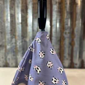 May include: A black gear shift knob with a lavender fabric cover. The fabric is patterned with white and black cow illustrations with pink accents. The gear shift cover is cone-shaped.