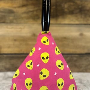 May include: A pink cone-shaped car gear shift knob with a black handle. The knob is covered in a fabric with a repeating pattern of yellow alien faces and white stars.