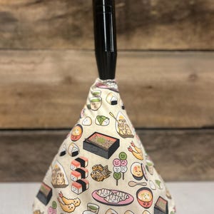 May include: A beige cone-shaped car gear shift knob with a black handle. The knob is covered in a fabric with a pattern of Japanese food, including sushi, dumplings, and tea.