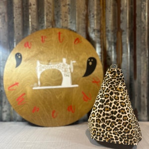 May include: A round wooden sign with the words "Sew Ready" written in red letters around the edge. A white sewing machine is in the center of the sign with two black ghost silhouettes on either side. A small leopard print fabric cone sits in front of the sign.